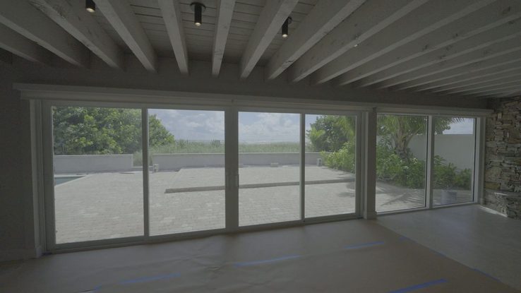 Family room with white beamed ceiling and sliding glass to the pool and sea grapes, roller shades raised