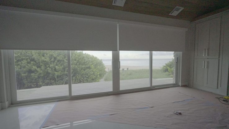 Primary suite bedroom with the SheerWeave 7200R Swan blackout shade raised, sliding glass open to the dune and the beach